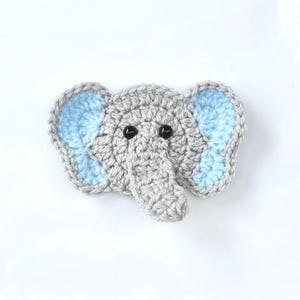 May include: A crocheted elephant applique in grey with light blue inner ears and black button eyes. The handmade elephant has a trunk and is suitable for baby items or other craft projects.