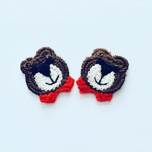 May include: Two brown crocheted bear faces with white eyes and black pupils. Each bear has a red crocheted bow tie.