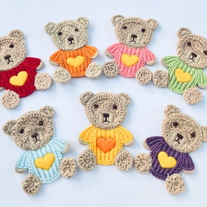 May include: Eight crocheted teddy bear appliques with embroidered hearts. The bears are beige with different coloured sweaters: red, orange, pink, green, light blue, yellow, purple.