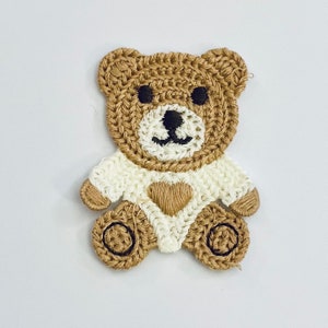 May include: A brown crocheted teddy bear applique with white accents. The bear has a heart-shaped chest and black embroidered eyes and nose.