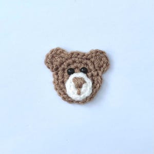 May include: A crocheted bear face applique in shades of brown and white. The bear has small black eyes and a white muzzle. The handmade item is likely for crafts, sewing, or embellishments.