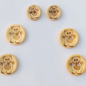 May include: Six wooden buttons featuring a teddy bear design. The buttons are beige and have a brown teddy bear holding a heart.