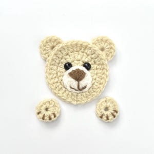 May include: A crocheted beige bear applique with black eyes, a brown nose, and white mouth. The bear has two separate crocheted beige paws.