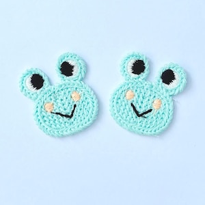 May include: Two light blue, knitted frog face patches. Each patch has black eyes, white eye highlights, peach-coloured cheeks, and a black smile. The patches are on a light blue background.