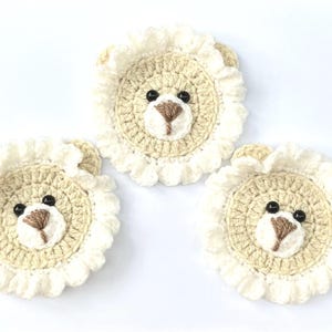 May include: Three handmade crochet lion coasters. Each coaster has a beige centre with a lion face, including black eyes and a brown nose. The lion's mane is a ruffled cream colour, and the ears are beige.