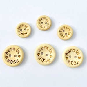 May include: Six round, light-coloured wooden buttons are displayed. Each button features two holes and is engraved with the words "Born in 2026" and small heart symbols. The buttons are arranged on a white surface.