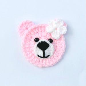 May include: A pink crocheted bear's head with a white flower. The bear has black bead eyes, a white muzzle, and a black nose and mouth. This handmade item is likely for a baby or child.
