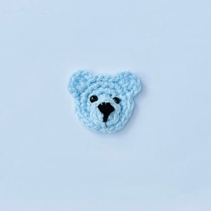 May include: A light blue crocheted bear face with black eyes and a black nose. The bear face is a handmade craft item, possibly a patch or applique for clothing or accessories. The bear's head is approximately 5 cm wide.