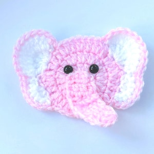 May include: A crocheted elephant head in pastel pink, featuring white inner ears and black button eyes. This handmade item is likely a decorative piece or a toy. The elephant's trunk is visible.