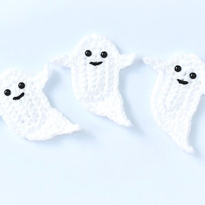May include: Three white crocheted ghosts with black eyes and smiling mouths. The ghosts are on a light blue background.
