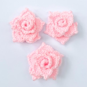 May include: Three individual light pink crochet flowers, each crafted with layered, spiralling petals. These handmade textile embellishments feature a soft, yarn-based texture and a dimensional, blooming rose-like shape with intricate stitching throughout.