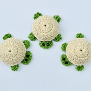 May include: Three crocheted turtle ornaments with green shells and black eyes. The turtles are made with beige yarn and have a simple design.