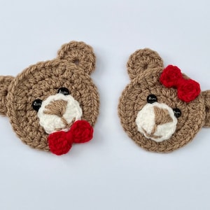 May include: Two crocheted brown teddy bear faces with black eyes. One bear has a red bow tie and the other has a red bow.
