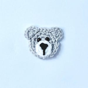 May include: A crocheted bear face applique in shades of grey, white, and black. The bear's head is grey, with white around the muzzle and black eyes and nose. The handmade item is likely for crafts or embellishments.