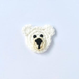 May include: A handmade, white crochet polar bear face with black button eyes and a black nose. The bear's snout is a light beige colour. The bear is centred on a white background.