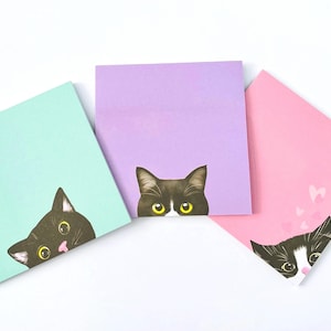May include: Three square sticky notes in pastel shades: pale blue, lilac, and pink. Each note has a black cat illustration peeking over the top. The pink note features small heart graphics. A fun stationery item.