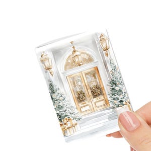 May include: A watercolor illustration of a white house with a gold door and two wreaths. The door has two windows and is flanked by two white trees with snow on them. There are two gold lanterns on the side of the house.