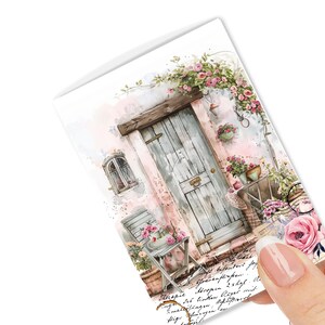 May include: A greeting card featuring a watercolor illustration of a rustic door adorned with climbing roses. The scene includes a small window, a chair, and potted flowers, all in soft pastel colors. The card is held by a hand.