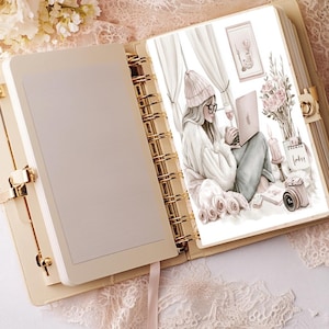 May include: An open planner with a gold spiral binding. The right page features a watercolour illustration of a woman wearing a pink beanie and a white sweater, sitting at a desk with a laptop and a glass of wine. The illustration is surrounded by pink flowers and a camera.