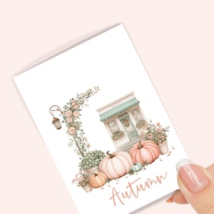 Op de afbeelding: Waterverfillustratie van een kaart met een herfsttafereel. De kaart toont een winkel met een groen luifel, pompoenen en bloemstukken. Het woord "Autumn" is geschreven in een scriptlettertype.