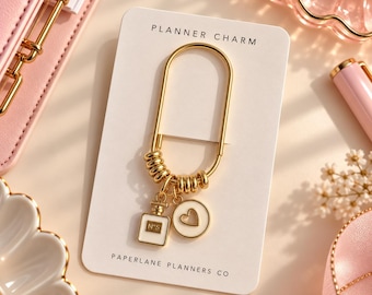 May include: A gold-toned metal paperclip-style planner charm attached to a white display card labeled PLANNER CHARM and PAPERLANE PLANNERS CO. The charm features gold rings and two white enamel pendants: a rectangular perfume bottle and a heart.