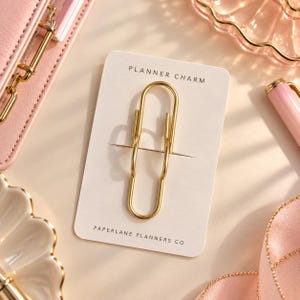 May include: A gold-toned metal paperclip charm attached to a rectangular white display card. The card features the text PLANNER CHARM at the top and PAPERLANE PLANNERS CO at the bottom in black sans-serif font.