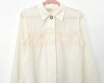 May include: Cream-colored long-sleeve blouse with a collared neckline and a decorative embroidered panel across the chest. The blouse features a single brown button closure and is hanging on a pink hanger.