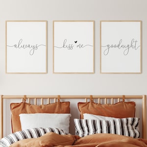 Op de afbeelding: Drie ingelijste prints met zwarte kalligrafie op een witte achtergrond. De prints lezen "always", "kiss me" met een klein hartje en "goodnight". De prints hangen aan een witte muur boven een houten bedframe met bruin beddengoed.