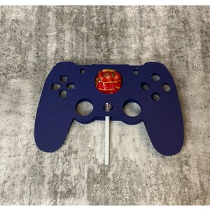 Puede incluir: Un soporte para piruletas de plástico azul con forma de mando de videojuego con una piruleta roja y blanca en su interior.