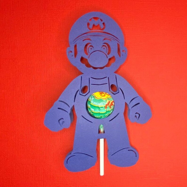 May include: Purple cardboard cutout of a video game character, Mario, holding a lollipop in his belly. The lollipop is green, yellow, and red.