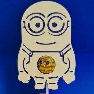 May include: Yellow Minion lollipop holder with a blue outline. The holder has a cut-out for a lollipop. The lollipop is yellow and brown with the word "CHOC" visible.