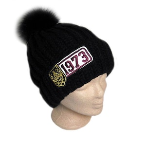 Op de afbeelding: Een zwarte gebreide muts met een grote, donzige zwarte pompon bovenop. De muts heeft een rechthoekige patch met het jaartal "1973" en een gouden embleem. De hoed wordt getoond op een mannequinhoofd tegen een witte achtergrond.