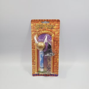 May include: A package of two Harry Potter themed stamp markers. The markers are in the shape of a golden snitch and a black hat. The package is on a white background.