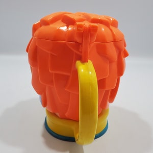 May include: Orange plastic cup with a yellow handle shaped like a cartoon character with orange hair. The cup has a blue base.