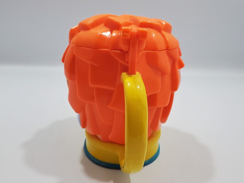 May include: Orange plastic cup with a yellow handle shaped like a cartoon character with orange hair. The cup has a blue base.