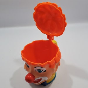 May include: Orange plastic clown head cup with a removable lid. The cup has a painted face with blue eyes, a red nose, and a yellow smile. The lid is also orange and has a wavy edge.