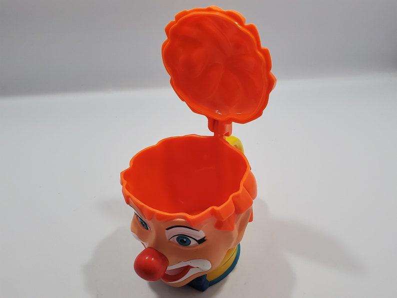 May include: Orange plastic clown head cup with a removable lid. The cup has a painted face with blue eyes, a red nose, and a yellow smile. The lid is also orange and has a wavy edge.