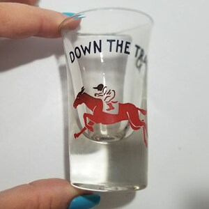 May include: A clear glass shot glass with a red and black design. The design features a horse and jockey with the text "DOWN THE TRACK" printed on the glass.