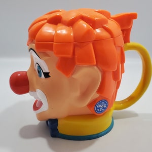 May include: Orange and yellow plastic mug shaped like a clown's head with a red nose and blue eyes. The mug has a handle and says "The Greatest Show on Earth" on the side.