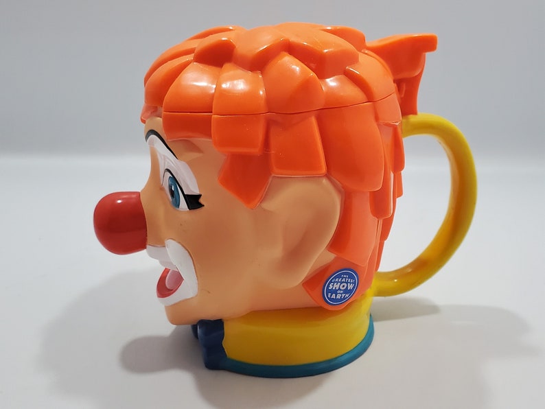 May include: Orange and yellow plastic mug shaped like a clown's head with a red nose and blue eyes. The mug has a handle and says "The Greatest Show on Earth" on the side.