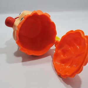 May include: Orange plastic toy with a clown face and a removable top. The toy is open and the inside is visible.