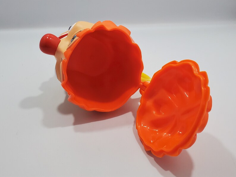 May include: Orange plastic toy with a clown face and a removable top. The toy is open and the inside is visible.