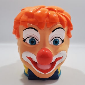 May include: A plastic clown head mug with orange hair, white makeup, blue eyes, and a red nose. The mug has a yellow and blue collar and a blue bow tie.
