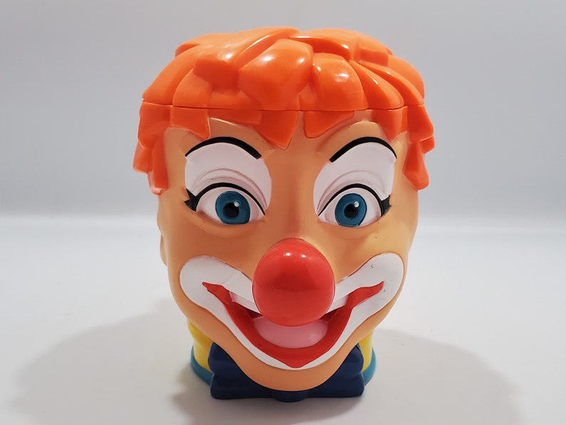 May include: A plastic clown head mug with orange hair, white makeup, blue eyes, and a red nose. The mug has a yellow and blue collar and a blue bow tie.