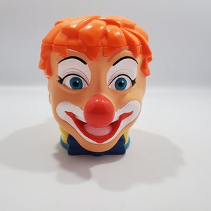 May include: A plastic clown head mug with orange hair, white makeup, a red nose, and a blue bow tie.