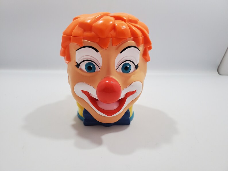 May include: A plastic clown head mug with orange hair, white makeup, a red nose, and a blue bow tie.