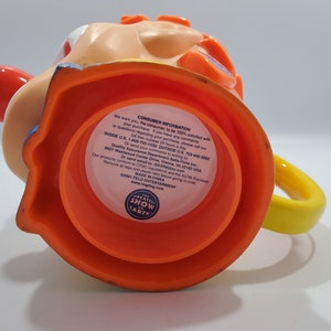 May include: Orange and yellow plastic mug shaped like a clown's head. The mug has a red nose, blue eyes, and a yellow handle. The bottom of the mug has a label that reads "The Greatest Show on Earth" and "Feld Entertainment".