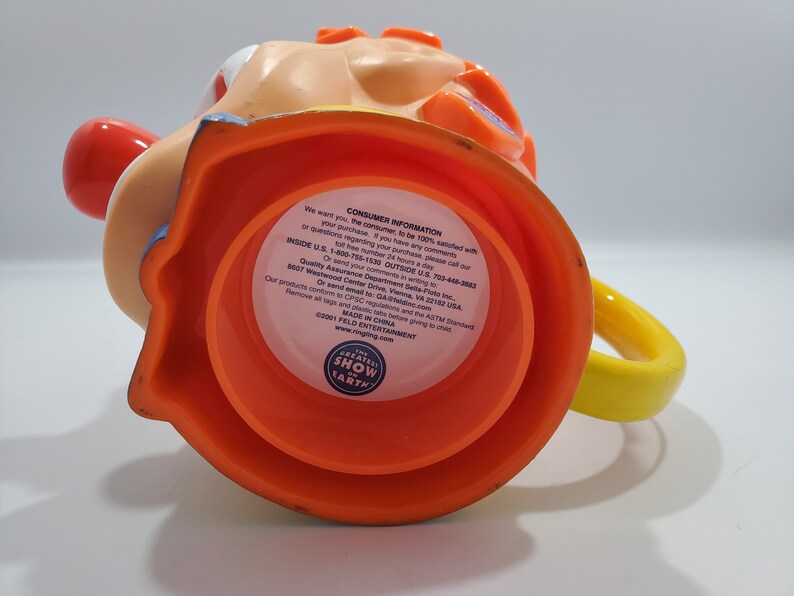 May include: Orange and yellow plastic mug shaped like a clown's head. The mug has a red nose, blue eyes, and a yellow handle. The bottom of the mug has a label that reads "The Greatest Show on Earth" and "Feld Entertainment".