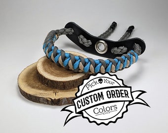 May include: A braided archery wrist sling in gray, blue, and tan paracord, with a black leather wrist strap. The sling is displayed on two wooden slices, with a custom order colors logo.