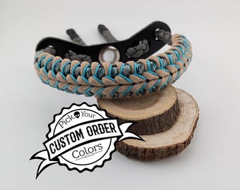 May include: A paracord bracelet featuring a braided design in tan, grey, and bright blue, attached to a black metal component with a central grommet. A white graphic overlay displays the text Pick Your CUSTOM ORDER Colors.